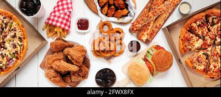 Tavola scena di assortiti take out o cibo di consegna. Pizza, hamburger, pollo fritto e contorni. Vista dall'alto verso il basso su uno sfondo di banner in legno bianco. Foto Stock