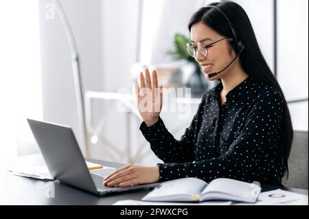 Amichevole bella donna asiatica intelligente indossare occhiali e cuffie, freelancer, dipendente di supporto o manager seduto al tavolo in ufficio che parla con il cliente o il dipendente, agitando la mano, sorridendo Foto Stock