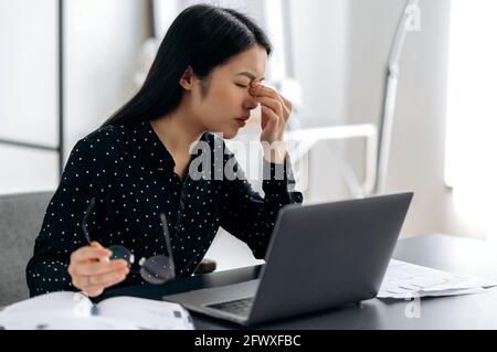 Overworked giovane donna asiatica triste, freelancer o manager, stanco di lavorare con il laptop, seduto sul posto di lavoro. Ragazza sconvolto sfregando il ponte nasale, tenendo gli occhiali in mano, chiude gli occhi Foto Stock