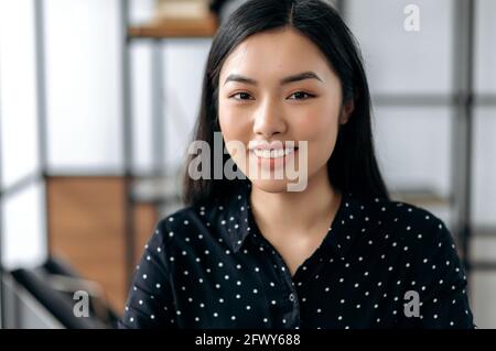 Primo piano ritratto di attraente cute successo sicuro giovane donna asiatica brunette, direttore d'ufficio, in formale camicia elegante, si siede contro sfondo offuscato ufficio, guarda la fotocamera, sorridente amichevole Foto Stock