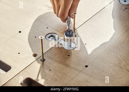 Un esperto artigiano assembla un grande tavolo da pranzo in legno girando il connettore del piano di lavoro con chiave all'aperto nelle giornate di sole, vista ravvicinata Foto Stock