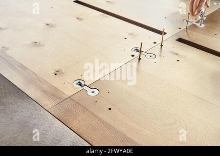 Un esperto artigiano assembla un grande tavolo da pranzo in legno girando il connettore del piano di lavoro con chiave all'aperto nelle giornate di sole, vista ravvicinata Foto Stock