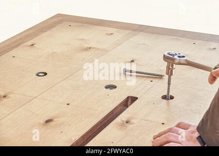 Artigiano esperto assembla grande tavolo da pranzo in legno girando bulloni con avvitatrice all'aperto nelle giornate di sole, vista ravvicinata Foto Stock