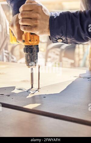 Il dipendente in giacca grigia assembla il nuovo tavolo da pranzo in legno tornando viti con cacciavite elettrico all'aperto nelle giornate di sole e closeup estremo Foto Stock