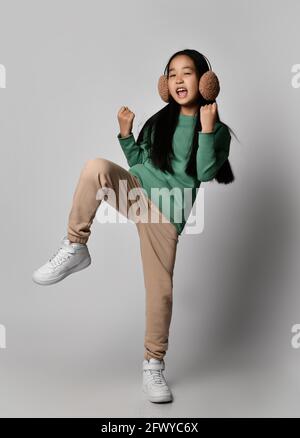 Bambina asiatica in cuffie verdi, abbigliamento sportivo e sneakers bianche si trova su una gamba che tiene il ginocchio in su e gesturing, ballando Foto Stock