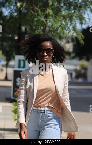 Modello abbagliante che cammina per strada con occhiali da sole. Primo piano ritratto, modello afroamericano, acconciatura naturale, borsetta sulla spalla, blazer casual Foto Stock