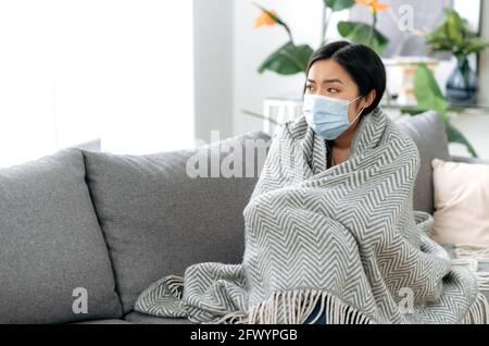 Il concetto di malattia e autoisolamento durante l'influenza o la covid19. Giovane donna asiatica stanca malata si siede turbata sul divano a casa, indossando una maschera medica protettiva, coperta di coperta, si guarda via tristemente Foto Stock