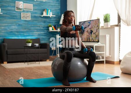 Donna africana che usa la sfera di stabilità che tiene le braccia estese che lavorano le spalle usando i manubri blu, nel soggiorno domestico per la formazione del muscolo e lo stile di vita sano, vestito in sportwear. Foto Stock