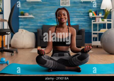 Ritratto di donna nera sorridente che pratica yoga a casa seduta in loto posa su mat mediting, pratica meditazione armonia della consapevolezza, persona afroamericana facendo allenamento rilassante. Foto Stock