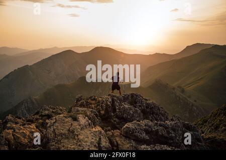 Vista posteriore dell'uomo che salpa su massi di alta cima di cresta di montagna sotto cielo nuvoloso al tramonto Foto Stock