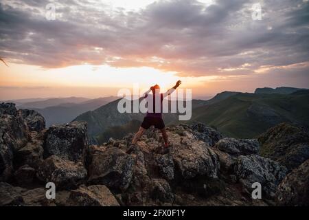 Vista posteriore dell'uomo che salpa su massi di alta cima di cresta di montagna sotto cielo nuvoloso al tramonto Foto Stock