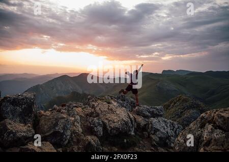Vista posteriore dell'uomo che salpa su massi di alta cima di cresta di montagna sotto cielo nuvoloso al tramonto Foto Stock