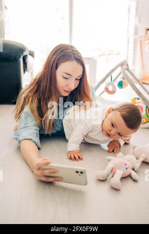 Donna giocosa che prende selfie con il bambino affascinante sul telefono cellulare mentre passa il tempo a casa Foto Stock