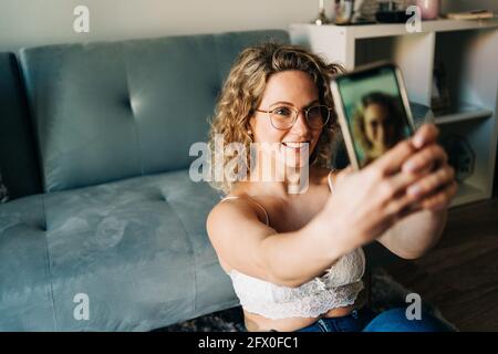 Dall'alto di auto assicurato giovane femmina millenario con riccia capelli biondi in pelle reggiseno un jeans seduto sul pavimento vicino al divano e prendendo selfie su intelligente Foto Stock