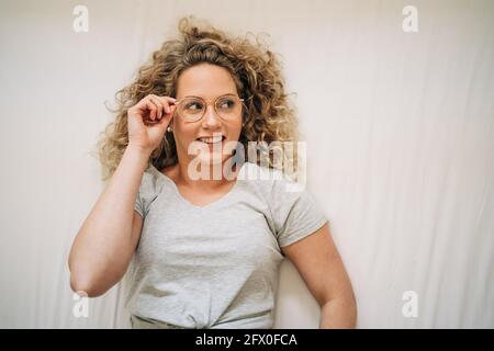 Vista dall'alto della giovane donna sicura con capelli biondi ricci in abiti casual regolare gli occhiali e sorridere mentre si sdraiava letto a casa Foto Stock