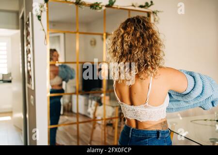 Vista posteriore PF giovane donna irriconoscibile con capelli biondi ricci in jeans e reggiseno in pizzo che indossa un caldo maglione in piedi in camera vicino a specchio nella giornata di sole Foto Stock