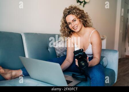 Allegro blogger giovane femmina con capelli biondi ricci casual outfit seduto sul divano e sorridente durante il trasferimento di foto da fotocamera professionale a. Foto Stock