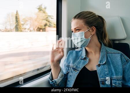 Giovane donna che indossa una maschera medica e saluto amico sulla stazione mentre si guida in treno e sorridendo Foto Stock