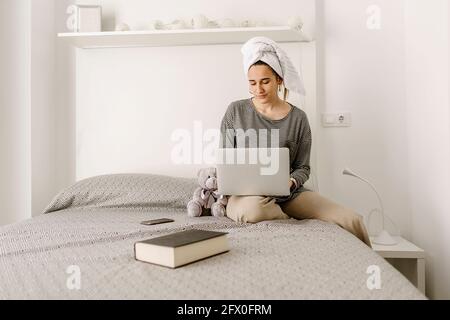 Giovane donna indipendente in abiti e asciugamano casual mentre si lavora a distanza sul netbook mentre si è seduti comodamente letto a casa Foto Stock