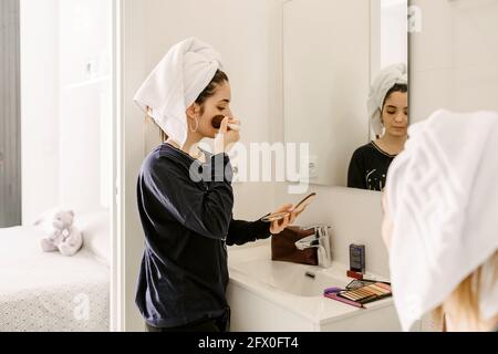 Vista laterale della giovane signora ispanica in abiti informali e. asciugamano sulla testa applicando il fondamento sul viso con la spazzola mentre in piedi davanti allo specchio vicino all'unr Foto Stock