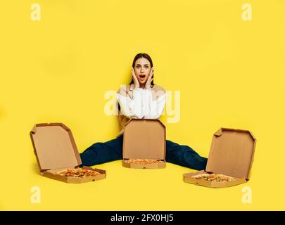 Emozionale sorpresa giovane donna circondata da tre pizze in scatole su sfondo giallo. Concetto di consegna della pizza. Foto Stock