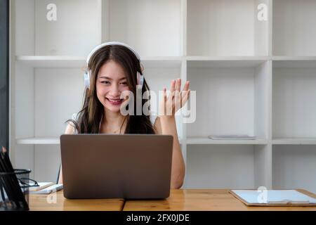 Vista laterale ripresa sorridente donna asiatica freelancer indossando cuffie, comunicando con il cliente tramite videochiamata al computer. Millennial piacevole professionale Foto Stock