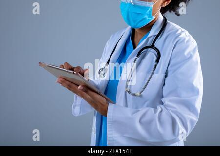 Sezione centrale del medico femminile che indossa la maschera facciale utilizzando il digitale tablet su sfondo grigio Foto Stock