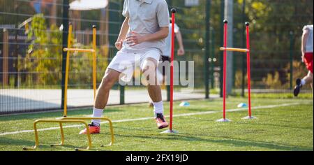 Giocatori di Teenage Football sul campo di allenamento. Young Boys Running Slalom traccia tra i poli di allenamento e saltando oltre gli ostacoli. Attrezzature da allenamento per il calcio Foto Stock