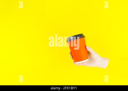 Tazza di carta arancione con tenuta a mano su sfondo giallo. Tazza da caffè di carta artigianale con spazio per la copia. Foto Stock