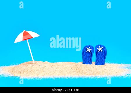 Ombrellone giocattolo e infradito nella sabbia su sfondo blu. Concetto creativo di vacanza in spiaggia. Foto Stock