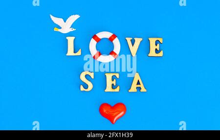 LOVE SEA composizione fatta di personaggi in legno, salvagente giocattolo, gabbiano e un cuore di plastica su sfondo blu. Sfondo estivo creativo. Foto Stock
