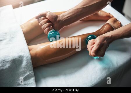 Bei piedi eleganti sdraiati sul tavolo da massaggio mentre il massaggiatore maschile fare massaggio con il bicchiere freddo in clinica Foto Stock