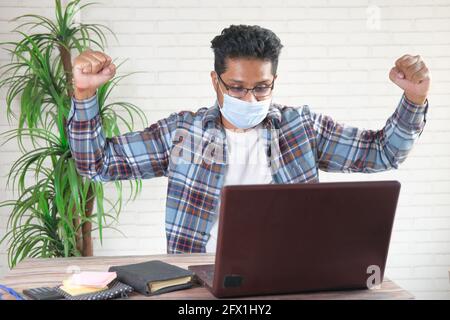 gioioso giovane uomo in maschera di faccia che lavora su un computer portatile Foto Stock