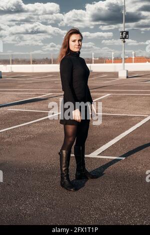 giovane donna bruna in vestiti neri e stivali in piedi a. parcheggio vuoto Foto Stock