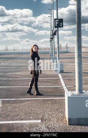 giovane donna bruna in vestiti neri e stivali in piedi a. parcheggio vuoto Foto Stock