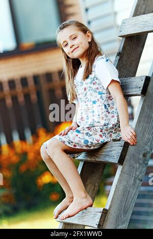 Una bella ragazza di campagna con i capelli sciolti in una semina estiva si siede su una scala di legno e guarda la macchina fotografica. Una giovane studentessa riposa in una giornata estiva nel villaggio. Foto Stock
