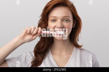 Giovane Donna spazzolando i denti usando lo spazzolino di plastica su sfondo grigio Foto Stock