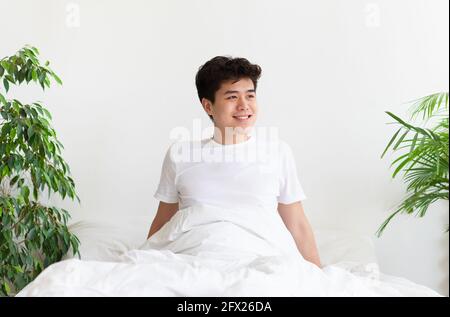 Felice giovane asiatico maschio si sveglia, si siede sul letto e guarda verso spazio vuoto Foto Stock