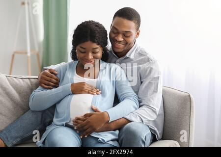 Sorridente uomo nero abbracciando la donna incinta, seduto sul divano Foto Stock