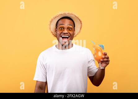 Uomo nero molto gioioso in abbigliamento casual che tiene un cocktail estivo rinfrescante, bocca di apertura in eccitazione su sfondo giallo Foto Stock