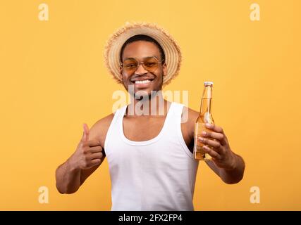Buon ragazzo nero che guarda nei vestiti estivi che tengono una bottiglia di birra e gesturing pollice su sfondo giallo studio Foto Stock