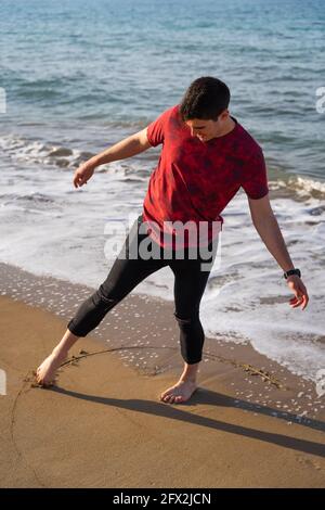 Uomo disegnando un cerchio con il piede sulla riva. È a piedi nudi e i suoi pantaloni sono arrotolati. È una giornata di sole e il mare è blu. È balanc Foto Stock