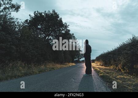 Un misterioso uomo incappucciato, tornato alla macchina fotografica. Guardando una lunga strada diritta, con fari per auto in lontananza. In una serata estiva. REGNO UNITO. Foto Stock