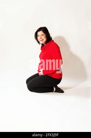 Giovane attraente donna incinta con il sorriso malizioso inginocchia e tiene per la pancia. Su sfondo bianco. Foto Stock