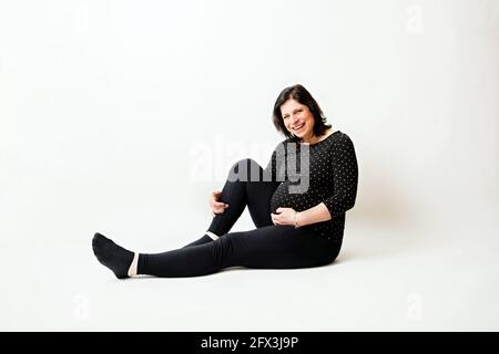 Felice e sorridente giovane donna incinta attraente, tenuta intorno al ventre, seduta su sfondo bianco. Foto Stock