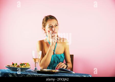 Giovane attraente donna intellettuale siede a tavola con vino, pasto e libro. Sta pensando. È interessata ai pensieri. Su sfondo rosa. Foto Stock