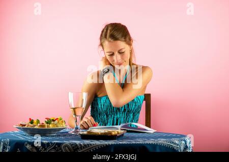 Bella giovane donna in abito blu si siede al tavolo con una piccola cena e un bicchiere di vino e legge un libro. Foto Stock