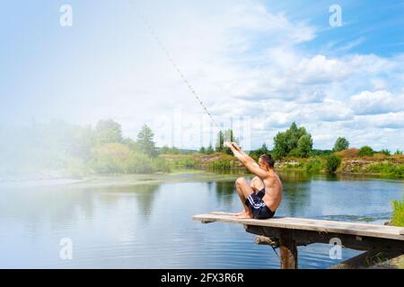 Ragazzo adolescente si siede su un ponte di legno che va a saltare nel fiume dalla corda oscillante nella soleggiata giornata estiva. Spazio di copia. Foto Stock