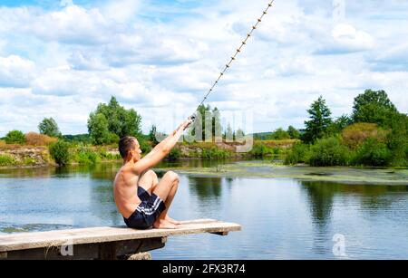 Ragazzo adolescente si siede su un ponte di legno che va a saltare nel fiume dalla corda oscillante nella soleggiata giornata estiva. Splendido paesaggio, spazio per la copia. Foto Stock