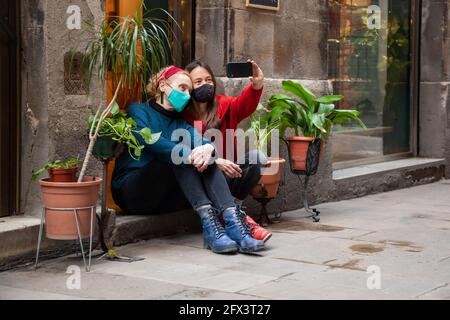 Due amici di donna che indossano maschere di protezione seduta vicino insieme prendendo selfie con smartphone. Concetto: Stile di vita cittadino, covid-19, tutti i giorni noi Foto Stock